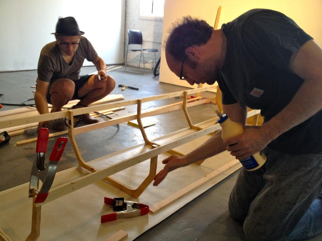 Two craftsmen measuring and assembling a long timber frame in a workshop